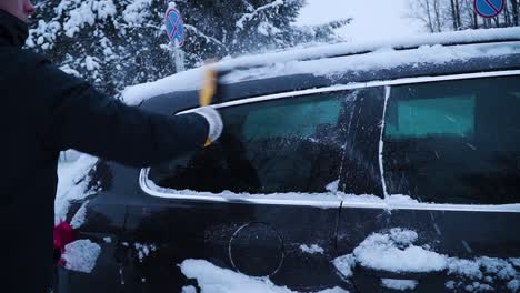 The-young-man-is-cleaning-car's-side-window-from-the-snow-with-a-yellow-brush-in-winter