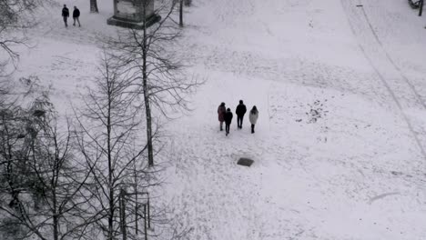 A-family-of-four-walks-leisurely-through-snow-covered-urban-park,-aerial-reveal