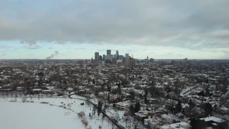Drone-Flies-Away-from-Minneapolis-Skyline,-Reveals-People-Skating-on-Pond-in-Winter