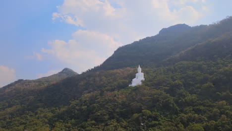 Luang-Por-Khao,-Wat-Theppitak-Punnaram,-aerial-4K-footage-towards-the-giant-White-Buddha-statue-on-a-mountain-side-in-Pak-Chong,-Thailand,-in-the-afternoon,-blue-cloudy-sky,-birds-flying-around