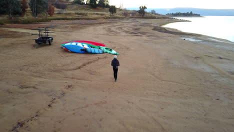 Lone-Man-Walking-On-Sandy-Shore-Passing-Through-Canoe-Boats-During-Daytime-In-Jindabyne,-New-South-Wales,-Australia