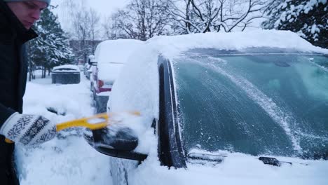 Young-man-is-cleaning-car's-front-and-side-windows-from-the-snow-with-yellow-brush-in-winter