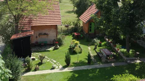Beautiful-small-village-household-with-2-houses-and-outdoors-toilet-in-the-garden-with-green-lawn-with-flowers-and-stone-path-surrounded-by-trees