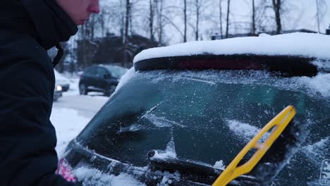 The-young-man-is-cleaning-car's-rear-window-from-the-snow-with-a-yellow-brush-in-winter