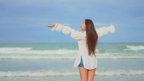Back-view-of-brunette-Asian-girl-on-seashore-raising-her-arms-to-the-sky