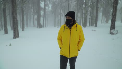 Front-view-following-a-man-walking-alone-through-a-snowy-forest,-while-wearing-a-bright-yellow-coat