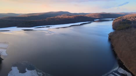 Aerial-drone-footage-of-a-partially-frozen-lake,-hills,-mountains,-and-forests-during-sunset-in-the-Appalachian-mountains-during-winter