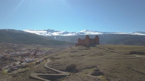 Aerial-ascending-shot-over-the-castle-of-La-Calahorra-on-top-of-a-hill-with-Sierra-Nevada-behind-during-the-winter