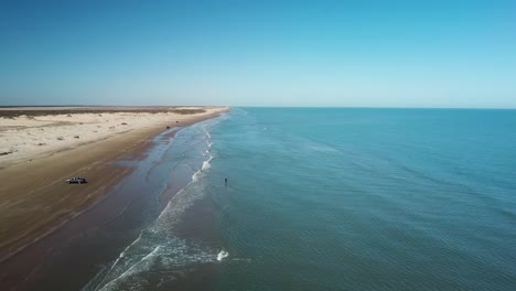 Aerial-drone-view-of-fisherman-standing-in-surf-on-beach-at-low-tide-on-a-gulf-coast-barrier-island-on-a-sunny-afternoon---South-Padre-Island,-Texas