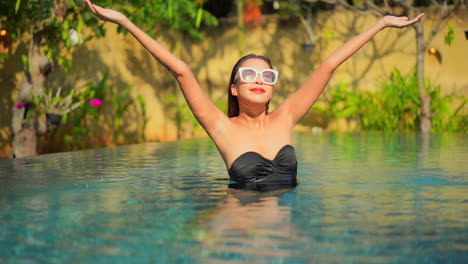 A-young-healthy-woman-in-a-resort-infinity-pool-raises-her-arms-to-greet-the-sun-and-day
