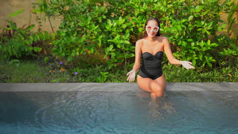 A-beautiful-fit-Asian-woman-in-a-black-bathing-suit-and-sunglasses-sits-on-the-edge-of-a-resort-pool-with-feet-in-the-water-together