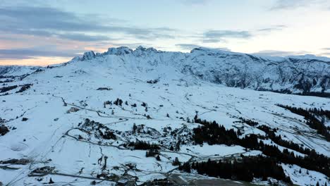 Panoramic-aerial-shot-of-mountain-range-in-the-Dolomites-covered-in-snow-seen-from-alpine-meadow-Seiser-Alm---Alpe-di-Siusi-plateau-in-South-Tyrol,-Italy-at-sunset