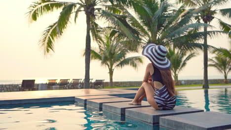 A-healthy,-fit,-woman-sitting,-with-her-back-to-the-camera,-on-the-edge-of-a-large-resort-swimming-pool-looking-out-at-the-ocean-horizon