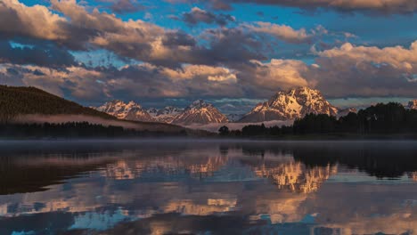 Beautiful-pink-and-golden-sunrise-at-reflective-mirror-lake-with-snow-capped-mountains-and-thick-clouds-flowing-by-in-Grand-Teton-National-Park,-Wyoming-United-States