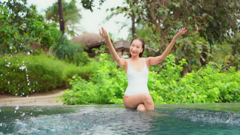 Fitte-Thailändische-Frau,-Die-Am-Rand-Des-Pools-Sitzt-Und-Mit-Ihren-Händen-In-Richtung-Der-Kamera-In-Einem-Exotischen-Tropischen-Resort-In-Einem-Grünen-Hintergrund-Schwimmbadwasser-Spritzt