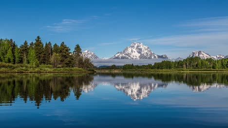 Time-lapse-of-clouds-flowing-along-huge-snow-capped-mountain-on-beautiful-reflective-mirror-lake-with-forest-pine-trees-in-Grand-Teton-National-Park,-Wyoming-United-States