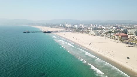 Sunny-sandy-Los-Angeles-beach-skyline-aerial-rising-towards-pier-above-turquoise-Pacific-ocean