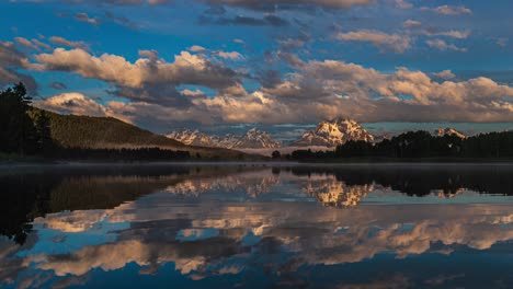 Wide-view-of-beautiful-pink-and-golden-sunrise-at-reflective-mirror-lake-with-snow-capped-mountains-and-thick-clouds-flowing-by-in-Grand-Teton-National-Park,-Wyoming-United-States