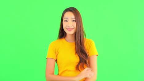 Schöne-Asiatische-Frau,-Die-Gelbes-T-shirt-Trägt,-Das-Die-Arme-Verschränkt-Und-Ok-geste-Mit-Zwei-Händen-Zeigt,-Die-Lächelnd-Zur-Kamera-Schauen,-Isoliert,-Studioaufnahme-Auf-Einem-Grünen-Studiohintergrund-Mit-Kopienraum-Für-Text