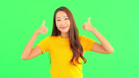 A-young-woman-against-a-solid-green-background-smiles-as-she-gives-the-thumbs-up-for-success