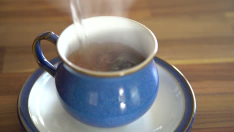 Heißes-Wasser,-Das-über-Einen-Teebeutel-Schießt,-Was-Zu-Einem-Dampfenden-Braunen-Tee-In-Einer-Blauen-Teetasse-Auf-Einem-Holztisch-Führt