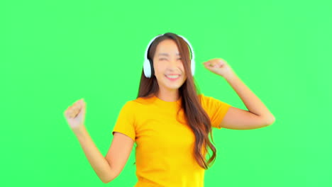 Young-Asian-Female-in-Front-of-Green-Screen-With-Wireless-Headphones-Dancing-in-Rthythm-of-the-Music,-Static-Slow-Motion