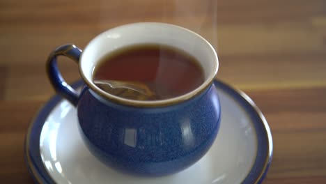 Small-bag-of-black-tea-bobbling-in-a-steaming-hot-water-in-a-blue-tea-cup-on-a-wooden-table-into-the-kitchen