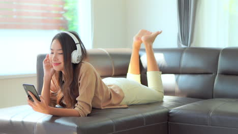 Smiling-young-woman-listens-music-with-headphones-lying-on-sofa-barefoot