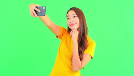 Lively-happy-Asian-woman-in-yellow-t-shirt-takes-selfies-with-phone-front-camera-showing-V-gesture-with-her-fingers