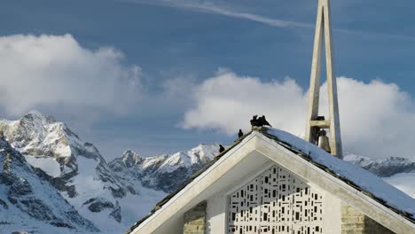 Gruppe-Schwarzer-Vögel,-Die-Auf-Einer-Kleinen-Kirche-Sitzen,-Die-Im-Schnee-Winterwunderland-Im-Zermatt-gletscher-skigebiet-In-Den-Schweizer-Alpen-Bedeckt-Ist