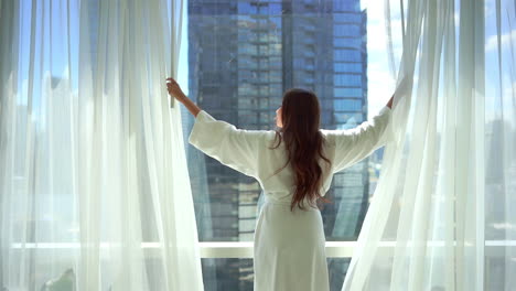 Back-view-of-woman-in-her-30th-wearing-bathrobe-opening-hotel-room-white-curtains-looking-through-window-enjoying-wellbeing,-unveiling-urban-city-view-on-skyscraper