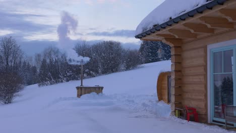 Smoking-furnace-at-dusk-in-winter-next-to-cabin-in-Poland,-wide-shot