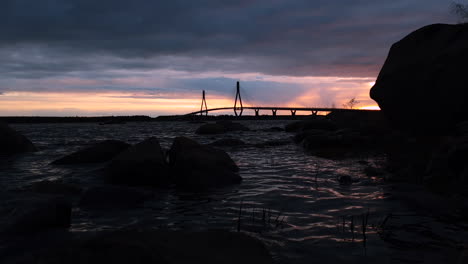 Dark-static-shot-of-distant-bridge-by-water,-dramatic-colorful-sunset