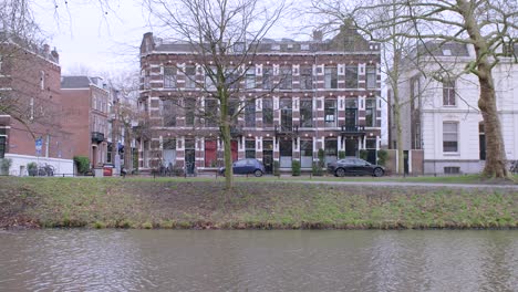 Un-Importante-Edificio-De-Ladrillo-De-Tres-Pisos,-Con-Tráfico-Circulando-Al-Frente,-Visto-Desde-El-Otro-Lado-Del-Canal-Que-Fluye-En-Utrecht,-Países-Bajos