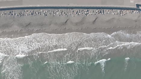 Static-birds-eye-aerial-of-waves-crashing-softly-on-the-sandy-shoreline-next-to-a-cement-seawall