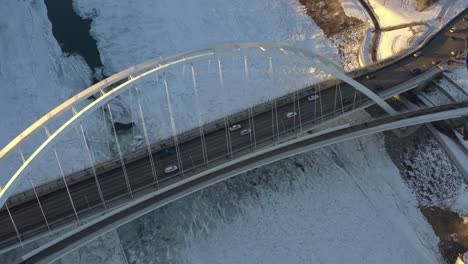 Aerial-drop-fly-over-Birds-Eye-View-dolly-roll-the-sunny-lit-white-walter-dale-modern-tie-arch-bridge-with-light-traffic-cracked-iced-North-Saskatchewan-River-snow-covered-futuristic-walkway-design