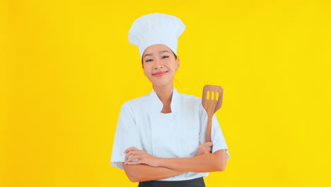 An-attractive-female-chef-in-a-traditional-chef's-uniform-is-standing-against-a-solid-background,-arms-folded,-wooden-spatula-in-hand