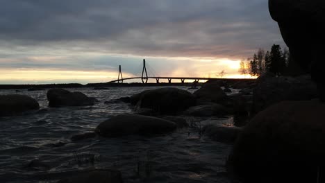Golden-sun-sets-behind-a-suspension-road-bridge-with-water-and-rocks-in-foreground