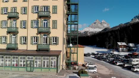 Descending-pedestal-aerial-shot-of-a-large-building-with-a-frozen-Misurina-Lake-and-Italy's-Three-Peaks-of-Lavaredo-in-the-distance