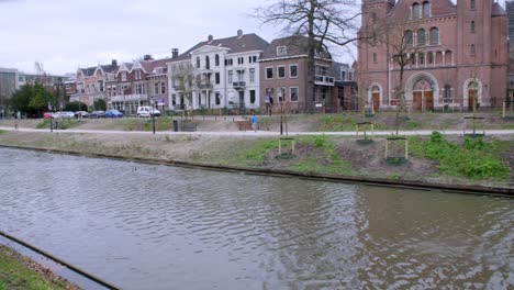 Un-Corredor-Solitario-Con-Top-Azul-Trota-A-Lo-Largo-De-Un-Camino-Junto-Al-Canal-En-Utrecht,-Países-Bajos,-Bordeado-De-árboles-Y-Viejos-Edificios-De-Ladrillo