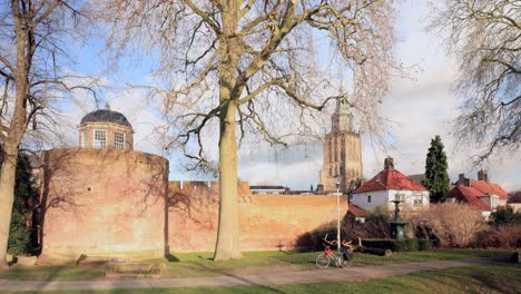 Time-lapse-of-historic-city-walls-with-park-and-stronghold-tower-in-winter-barren-landscape-with-Walburgis-church-tower-of-Zutphen,-The-Netherlands,-in-the-background-against-a-blue-sky-with-clouds