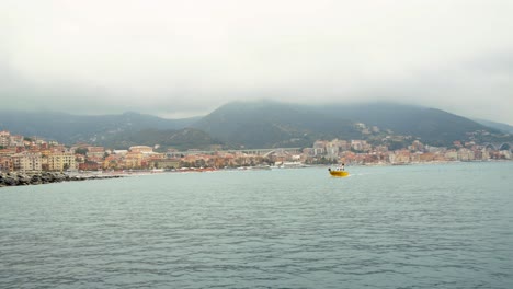 Small-yellow-boat-sails-in-the-bay-of-the-port-of-Varazze-on-a-cloudy-day,Background-Varazze-town-,-Liguria-Italy
