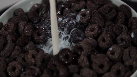 Milch-Wird-In-Die-Schüssel-Mit-Schokoladenmüsli-Gegossen,-Milchtropfen-Fallen-Auf-Das-Kakaomüslifrühstück,-Nahaufnahme,-Weißer-Hintergrund