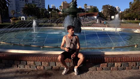 A-man-sits-on-the-edge-of-a-fountain-and-enjoys-a-coffee-as-he-relaxes-in-the-park-in-Montevideo,-Uruguay