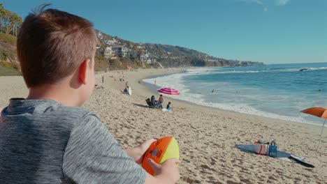 Boy-observing-waves-crashing-on-beach,-holding-a-football