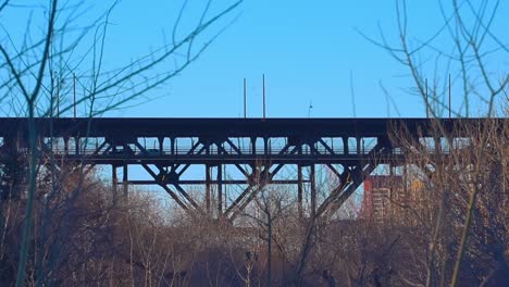shadow-silhouette-of-cars-crossing-the-high-level-steel-truss-bridge-vew-from-the-Kinsmen-park-and-looking-upwards-behind-bare-branched-trees-bushes-with-buildings-behind-vintage-historic-overpass