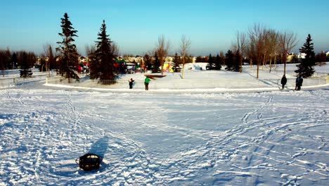 Aerial-winter-rise-fly-over-hay-stacks-covered-in-snow-picnic-tables-next-to-iron-fire-pits-with-optional-grills-surrounding-a-man-made-skating-rink-connected-to-an-ice-track-around-a-playground-1-2