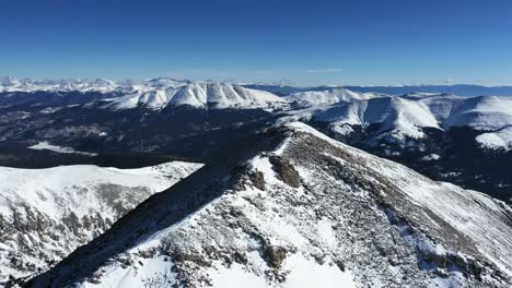 Aerial-View,-Person-on-Mountain-Peak-and-Snow-Capped-Range-on-Sunny-Winter-Day