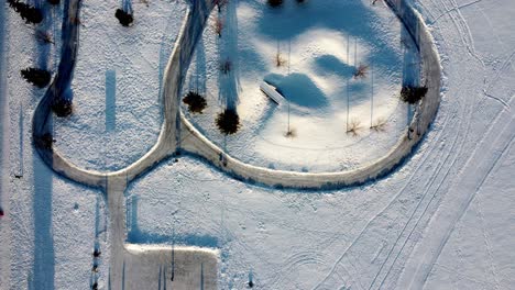 Aerial-birds-eye-view-overlooking-snow-covered-winter-manmade-dual-fish-oval-skating-track-with-a-slide-hill-for-children-to-go-sledding-and-ice-path-links-to-a-rectangular-hockey-rink-tall-pines-1-3