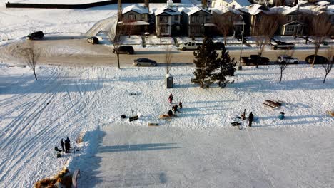 winter-aerial-rise-over-residential-community-manmade-ice-rink-people-practicing-skating-hockey-and-learning-to-skate-community-park-out-house-people-communing-at-picnic-table-fire-pit-hay-stacks-1-3